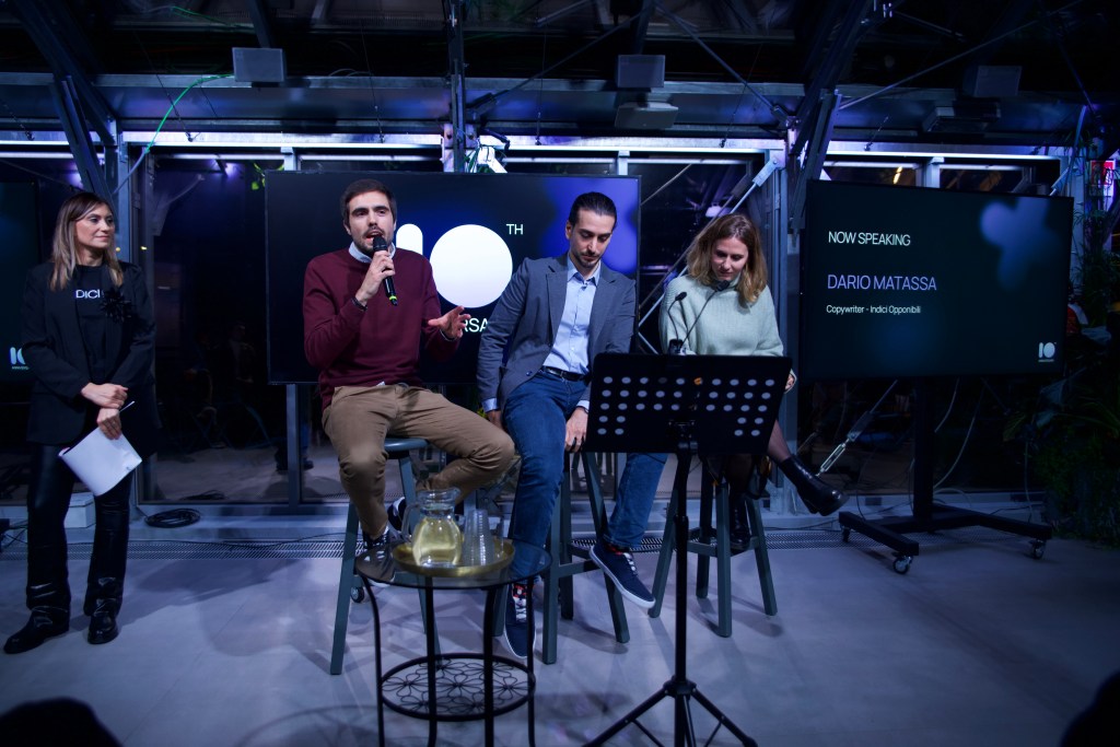 Panel talk dell’evento Indici10 con Dario Matassa sul palco insieme ad altri ospiti, all’interno di uno spazio contemporaneo illuminato da schermi digitali e luci scenografiche.