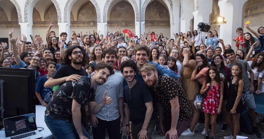 Foto di gruppo del team di Space Valley insieme al pubblico durante un evento dal vivo all’interno di uno spazio storico affollato di ragazzi e creator.