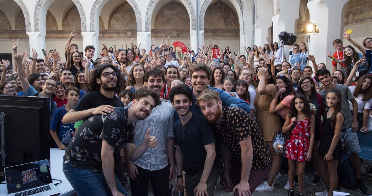 Foto di gruppo del team di Space Valley insieme al pubblico durante un evento dal vivo all’interno di uno spazio storico affollato di ragazzi e creator.